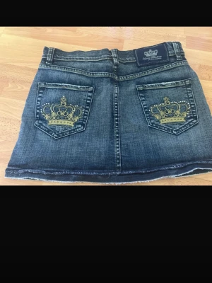 Jeanskjol S/M  med guldbroderi från Victoria Beckham  - Cool jeanskjol i mörkblå denim från Victoria Beckham med två bakfickor prydda med guldfärgade kronbroderier. Rå kant nertill för en edgy look. Perfekt för dig som vill sticka ut med detaljer.