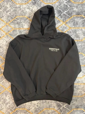 Svart Essentials Fear of God hoodie - Snygg svart hoodie från Essentials Fear of God med vit logga tryckt på bröstet. Klassisk oversized passform med stor huva och ribbade muddar vid ärmar och midja. Perfekt för en avslappnad streetwear-look.