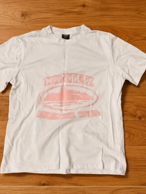 Vit t-shirt med rosa tryck - Vit t-shirt i bomull med ett blekt rosa tryck framtill. Klassisk rund hals och korta ärmar. Perfekt för en avslappnad och enkel stil. Trycket ger en vintagekänsla och t-shirten har en normal passform.