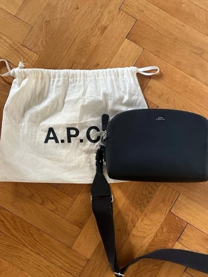 Svart axelväska från A.P.C. - Snygg svart axelväska från A.P.C. i slätt läder med minimalistisk design och silverfärgade detaljer. Väskan har en bred justerbar axelrem i textil och har två fack som stängs med dragkedja. Väskan har även ett mindre fack i ett ut av dem större facken och har även en liten ficka på baksidan. Perfekt för dig som gillar stilrena accessoarer. Utmärkt för både herr och dam! (Denna modell är en limited edition) bredd:24cm, höjd:17cm, djup:8cm