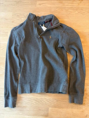 Grå halvzip tröja från Polo Ralph Lauren - Mysig grå långärmad tröja från Polo Ralph Lauren med halvzip och liten broderad logga på bröstet. Tröjan har hög krage och ribbade muddar vid ärmslut och nederkant. Perfekt för lager på lager och enkel att matcha.