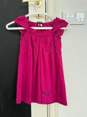 Rosa kortklänning med volang - Supersöt rosa kortklänning med volangdetaljer och smock upptill. Klänningen är ärmlös och har en loose passform som ger en avslappnad vibe. Tillverkad i mjuk bomull, perfekt för varma dagar. Märket Only Style syns broderat längst ner på framsidan.