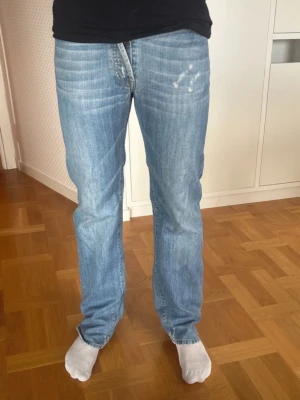 Ljusblå raka jeans - Säljer ett par ljusblå jeans med rak passform och klassisk femficksdesign. Jeansen har slitningar på låret för en avslappnad look och är tillverkade i denim. Perfekta för dig som gillar en enkel och tidlös stil.