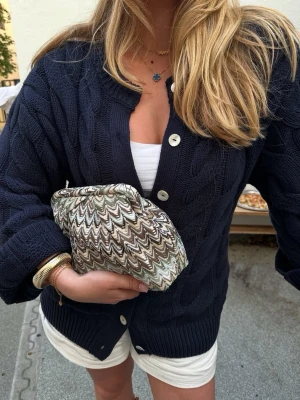 Mörkblå kabelstickad kofta - Snygg mörkblå kabelstickad kofta med stora vita knappar framtill. Koftan har en avslappnad passform, ribbade muddar och rund halsringning utan krage. Perfekt att slänga över en topp för en chill look.