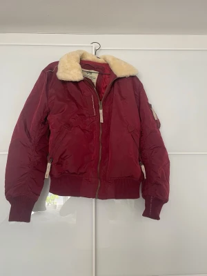 Vinröd bomberjacka Alpha Industries - Snygg vinröd bomberjacka från Alpha Industries med krage i beige fuskpäls. Jackan har dragkedja framtill, ribbade muddar och fickor med dragkedja. Klassisk pilotstil och perfekt för kyligare dagar.