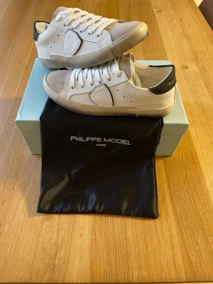 Philippe Model vita sneakers med mockadetaljer - Snygga vita sneakers från Philippe Model med beige mockadetaljer och svart hälparti. Skorna har klassisk låg profil, vita skosnören och ikonisk sköld på sidan. Tillverkade i en mix av läder och mocka för en exklusiv känsla.