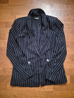 Svart randig kavaj från Noisy May - Snygg svart kavaj med vita vertikala ränder från Noisy May. Klassisk passform med smal krage och två fickor framtill med dekorativa detaljer. Tunnare material och helfodrad för en clean look.