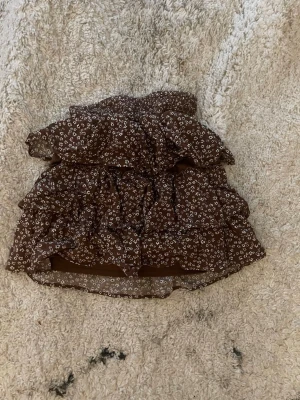 Brun volangkjol med blommönster - Supersöt brun kortkjol med små vita blommor och flera volanger. Kjolen har en luftig och flowy känsla, perfekt för varmare dagar. Elastisk midja för skön passform och trendig look. Barn storlek men väldigt stretchig
