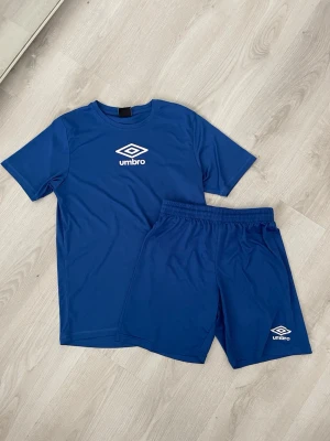 Blått träningsset från Umbro - Säljer ett blått träningsset från Umbro med t-shirt och shorts. Setet är gjort i ett lätt och snabbtorkande material, perfekt för träning eller fotboll. Både tröjan och shortsen har Umbro-loggan tryckt i vitt framtill.