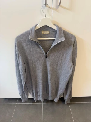 Lorenzo Magani Merinoull 1/4 Zip Made in Italy, stl L - Snygg grå stickad tröja från Lorenzo Magani med halvzip och hög krage. Tröjan har långa ärmar, ribbade muddar och är tillverkad i mjukt material som passar perfekt för kyligare dagar.