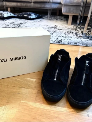 Svarta sneakers från Axel Arigato - Snygga svarta sneakers från Axel Arigato i mocka och läder. Skorna har dragkedja framtill istället för klassiska snören och en chunky sula. Perfekta för dig som gillar stilrena och moderna detaljer. OBS lite trasiga i hläen men inget man märker eller känner av