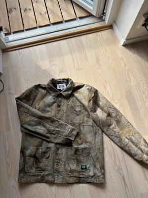 Camo overshirt med lövprint - Säljer en overshirt i camomönster med löv och naturmotiv. Skjortan har krage, knappstängning framtill och två stora fickor nedtill. Materialet känns slitstarkt och perfekt för lager-på-lager. Perfekt för dig som gillar streetwear och naturinspirerad stil.
