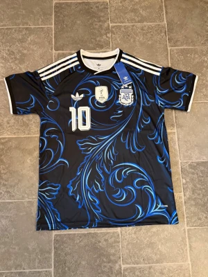 Argentina Messi tröja svart/blå Adidas - Snygg Argentina fotbollströja från Adidas med Messi och nummer 10. Tröjan är svart med blåa mönster, vita detaljer och tre vita ränder på axlarna. FIFA-märke och AFA-logga på bröstet. Perfekt för fotbollsfans!