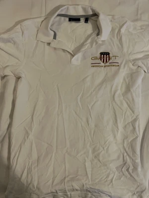 Vit pikétröja från Gant - Klassisk vit pikétröja från Gant med broderad logga och texten 'American Sportswear' på bröstet. Tröjan har krage, korta ärmar och knappar vid halsen. Perfekt för en clean och sportig stil.