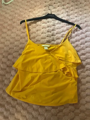 Gul volang topp med smala band - Säljer en solgul linnetopp med smala axelband och volangdetaljer framtill. Toppen har en omlottkänsla och är perfekt för varma dagar. Lätt och luftig modell som ger en fräsch look.