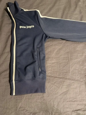 Mörkblå ziptröja från Palm Angels - Snygg mörkblå ziptröja från Palm Angels med vit dragkedja och vita detaljer längs ärmarna. Tröjan har hög krage, fickor med dragkedja och logga på bröstet. Tillverkad i polyester med elastan i muddarna för extra komfort.