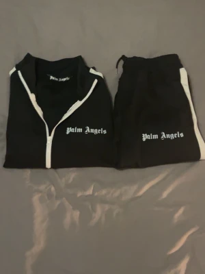 Palm Angels dress Svart - Säljer ett svart träningsset från Palm Angels med vita detaljer. Setet består av en zip-jacka med vita ränder längs ärmarna och dragkedja framtill samt matchande byxor med vita sidoränder. Båda delarna har Palm Angels-loggan tryckt i vitt. Perfekt för sport eller chill.