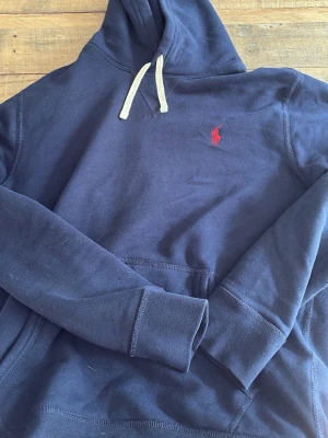 Marinblå hoodie från Polo Ralph Lauren - Snygg marinblå hoodie från Polo Ralph Lauren med klassisk röd logga broderad på bröstet. Hoodien har vit dragsko, känguruficka och är tillverkad i mjuk bomullsblandning. Perfekt för en chill och stilren look.