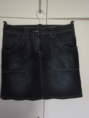 Mörkblå jeanskjol - Snygg mörkblå jeanskjol i kort modell med stora framfickor och två bakfickor med lock och knapp. Kjolen har bälteshällor och klassisk jeansknäppning framtill. Perfekt till en avslappnad och trendig stil. Den perfekta jeanskjolen att ha nu i höst tillsammans med en vinröd långärmad!🍂🍁