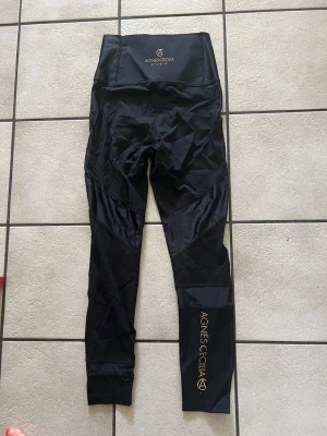 Svarta leggings Agnes Cecilia Studio storlek 36/38 - Snygga svarta leggings från Agnes Cecilia Studio med hög midja och guldfärgad logga både fram och på benet. Tillverkade i stretchigt material som sitter skönt och följsamt. Perfekta för träning eller chill.