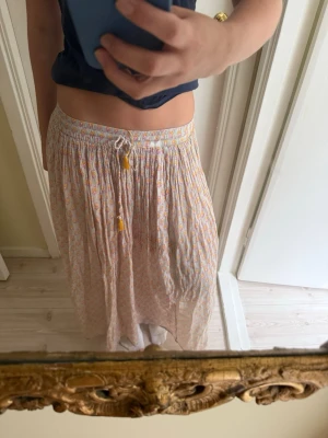 Långkjol med blommigt mönster och snörning - Luftig långkjol i ljusa toner med småblommigt mönster i rosa och gult. Kjolen har en bekväm resårmidja med snörning och tofsar framtill. Perfekt för varma dagar och ger en avslappnad boho-vibe. Denna kjol är från en affär i Kenya. 