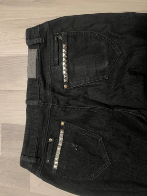 Svarta bootcut jeans med slits - Säljer ett par svarta bootcut jeans med slits nertill på benen och mörkblå kil. Klassisk femficksmodell med knapp och dragkedja. Jeansen har en cool och unik look som sticker ut. Perfekta för dig som vill ha något annorlunda i garderoben.