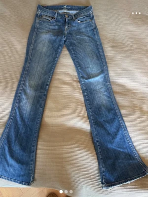 Blå bootcut jeans med slitningar från 7 for al mankind - Säljer ett par blå bootcut jeans med snygga slitningar och klassisk femficksmodell. Jeansen har en lätt tvättad look och vida ben som ger en retrovibe. Perfekta för dig som gillar en avslappnad men trendig stil.