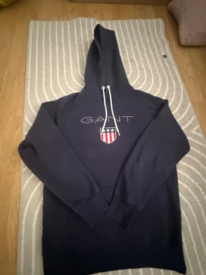 Mörkblå hoodie från GANT - Mörkblå hoodie från GANT med broderad logga och sköld på bröstet. Klassisk modell med huva och vita dragsnören. Perfekt för en avslappnad och stilren look.
