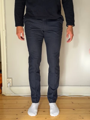 Mörkblå kritstrecksrandiga kostymbyxor från Riley - Mörkblå kritstrecksrandiga kostymbyxor från Riley i storlek 150 • Passform: Slim fit / Modern passform • Skick: Mycket gott skick Klassiska och stilrena kritstrecksrandiga byxor med pressveck och fin struktur. Perfekta till både vardag och mer uppklädda tillfällen. Sitter snyggt längs benen med avsmalnande benslut och har fickor fram samt bak. 