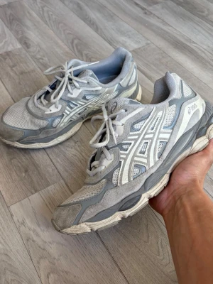 Asics Gel-Nyc - Snygga gråa sneakers från Asics med vita och ljusgrå detaljer. Skorna har en sportig design med meshpaneler för bra ventilation och klassiska Asics-loggor på sidorna. Tjock sula för extra komfort och snörning framtill. Perfekta för en avslappnad streetwear-look.