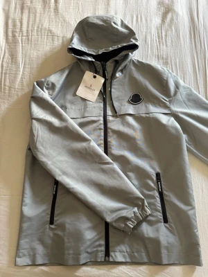 Ljusgrå vindjacka med huva Moncler - Snygg ljusgrå vindjacka från Moncler med svart dragkedja, två fickor med dragkedja och elastiska muddar. Jackan har huva och ikonisk Moncler-logga på bröstet. Insidan har ett färgglatt serietryck. Perfekt för blåsiga dagar.
