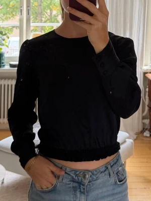 Svart långärmad blus från Bikbok - Snygg svart långärmad topp med croppad passform och rund hals. Toppen har resår i midjan och knappar vid ärmsluten för en extra detalj. 