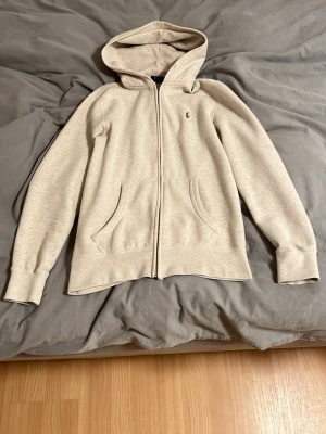 Beige hoodie från Polo Ralph Lauren - Snygg beige hoodie från Polo Ralph Lauren med dragkedja framtill och två fickor. Klassisk broderad logga på bröstet och mjukt material som känns skönt mot huden. Perfekt för en avslappnad och stilren look.