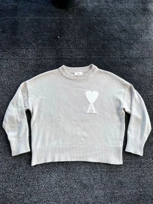 Ami Paris stickad - Snygg beige stickad tröja med rund hals och ribbade muddar. På bröstet finns en vit hjärtdetalj med bokstaven A. Tröjan har en avslappnad passform och är perfekt för lager på lager. Materialet känns mjukt och bekvämt. Storlek M men passar S, skick 9/10, äkta! Pris går att diskutera✅