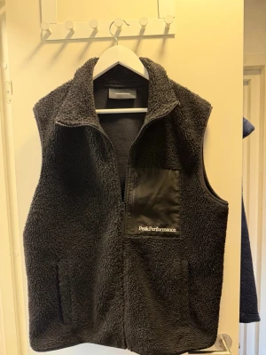 Svart fleeceväst Peak Performance - Säljer en svart fleeceväst från Peak Performance med dragkedja framtill och en bröstficka i nylon med logga. Västen har hög krage och två sidofickor. Perfekt att slänga över en hoodie för extra värme och stil.   Storlek XL men passar L