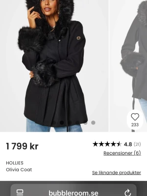 Svart kappa med fuskpäls från Hollies - Stilren svart kappa från Hollies med stora detaljer i svart fuskpäls på huva och ärmslut. Kappan har en feminin siluett, knäppning framtill och diskret logga på ärmen. Perfekt för dig som vill ha en trendig och mysig look under kalla dagar.