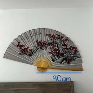 Stor dekorativ solfjäder 90 cm - Säljer min fina älskade väggsolfjäder pga att den inte passar i min nya lägenhet🥲, ca 90 cm bred (markerad på bilden) , med grå bakgrund och handmålat blommotiv i rött, svart och lila. Solfjädern har trähandtag av bambu och är perfekt som unik väggdekoration. Pris kan diskuteras eftersom att jag haft den på väggen ett tag.