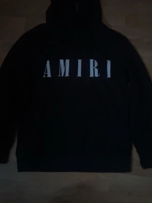 Amiri  - Snygg svart hoodie från Amiri med stor vit logga över bröstet. Klassisk passform med huva och långa ärmar. Perfekt för dig som gillar streetwear och vill ha en stilren look. Tillverkad i mjukt material för extra komfort.