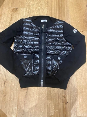 Svart pufferjacka från Moncler - Snygg svart pufferjacka från Moncler med glansig vadderad front och två fickor med knappar. Jackan har ribbade muddar, dragkedja framtill och en patch med Moncler-logga på ärmen. Perfekt för kalla vinterdagar och riktigt trendig look.
