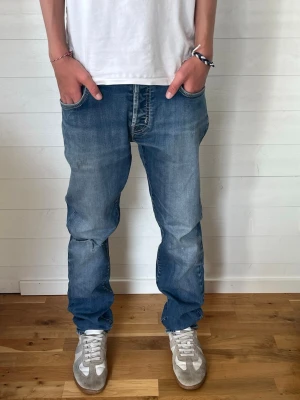 Blå raka jeans med slitningar - Säljer ett par blå jeans med raka ben och lätt slitna detaljer på knäna. Jeansen har klassisk femficksdesign och normal midja. Materialet är denim i bomull och passformen är avslappnad men ändå snyggt åtsittande. 