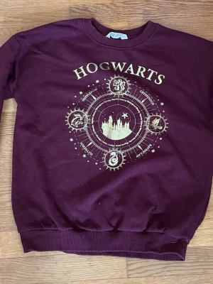 Vinröd Hogwarts sweatshirt från H&M - Vinröd sweatshirt från H&M med stort Hogwarts-tryck i guld på bröstet. Tröjan har ribbade muddar vid ärmslut och nederkant samt rund hals. Perfekt för alla Harry Potter-fans som vill ha något bekvämt och snyggt.