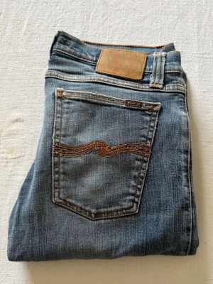 Blå jeans från Nudie Jeans - Säljer ett par klassiska blå jeans från Nudie Jeans med raka ben och snygga kontrastsömmar. Jeansen har två bakfickor med den ikoniska vågiga sömmen och är gjorda i mjuk denim. Perfekta för dig som gillar en tidlös och avslappnad stil. Storlek W32 L34
