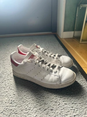 Vita Adidas Stan Smith sneakers - Klassiska vita Adidas Stan Smith sneakers med röda detaljer på hälen och tungan. Skorna har lågt skaft, perforerade ränder på sidorna och är tillverkade i slätt läder. Perfekta för en clean och tidlös stil.