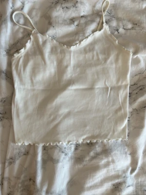 Vit ribbad linnetopp med volangkant - Säljer en söt vit linnetopp med tunna axelband och volangkant längst ner. Toppen är ribbad och har en croppad passform som passar perfekt till höga jeans eller kjol. Enkel och stilren design som är lätt att matcha.