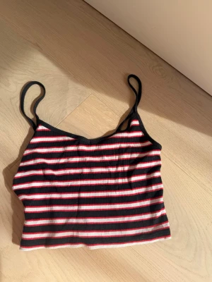 Randig ribbad linnetopp - Säljer en croppad linnetopp med smala axelband och ribbad struktur. Toppen har svarta, vita och röda horisontella ränder och är perfekt för varma dagar. Enkel och snygg att matcha med jeans eller kjol.