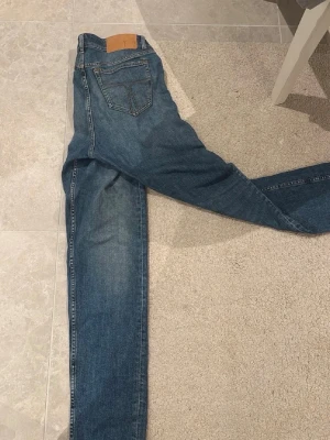 Blå raka jeans i modell REX - Säljer ett par klassiska blå jeans i modellen REX med raka ben och normalhög midja. Jeansen har fem fickor, kontrastsömmar och en patch bak i midjan. Tillverkade i denim med snygg tvätt och slitningar. Perfekta till en avslappnad stil.