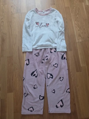 Mysig pyjamas med hjärtmönster - Supersöt pyjamas med långärmad vit tröja och rosa detaljer vid halsen. Tröjan har broderad text 'love' och små hjärtan framtill. Byxorna är ljusrosa med svarta och rosa hjärtan över hela. Tillverkad i mjukt, fluffigt material som är perfekt för mysiga kvällar.