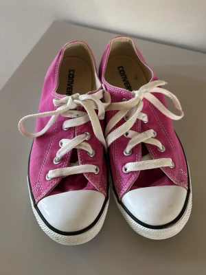 Rosa Converse låga sneakers - Säljer ett par klassiska Converse sneakers i en snygg rosa färg med vita gummisulor och tåhättor. Skorna har vita skosnören och silverfärgade öljetter. Tillverkade i canvasmaterial och har den ikoniska Converse-looken. Perfekta för en avslappnad och trendig stil.