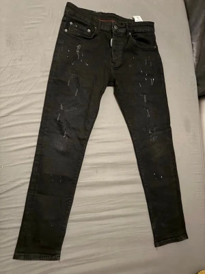 Svarta skinny jeans med slitningar - Säljer ett par svarta  jeans med slitningar och små färgstänk framtill. Jeansen har fem fickor, knappgylf och en smal passform. Materialet ser ut att vara stretchigt och bekvämt, perfekt för en trendig streetwear-look.
