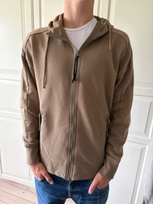 Beige/brun CP Company hoodie med dragkedja  - Säljer en beige/brun CP Company hoodie med dragkedja och coola glasögon-detaljer på huvan. Hoodien har två fickor med dragkedja och justerbara snören i huvan. Perfekt för dig som vill sticka ut lite extra. Tillverkad i mjukt bomullsmaterial. Tröjan är i storlek L men passar även M. Hör av er vid frågor och priset är inte hugget i sten!